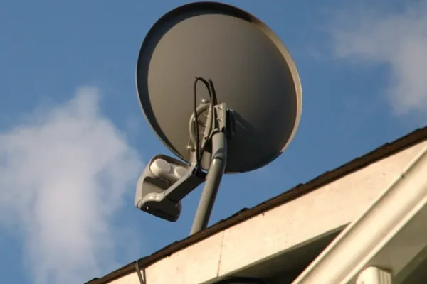 A gray satellite dish mounted on the edge of a light-colored roof, set against a bright blue sky with white clouds.